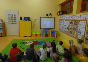 Grupa dzieci siedzi na dywanie przed tablicą interaktywną i ogląda prezentację multimedialną.